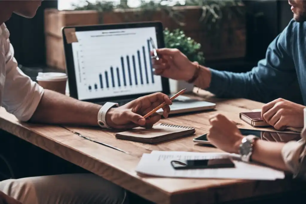 Two people sit at a wooden table with notebooks, a laptop displaying a rising bar graph, and documents, discussing and pointing at the screen in a collaborative business meeting.