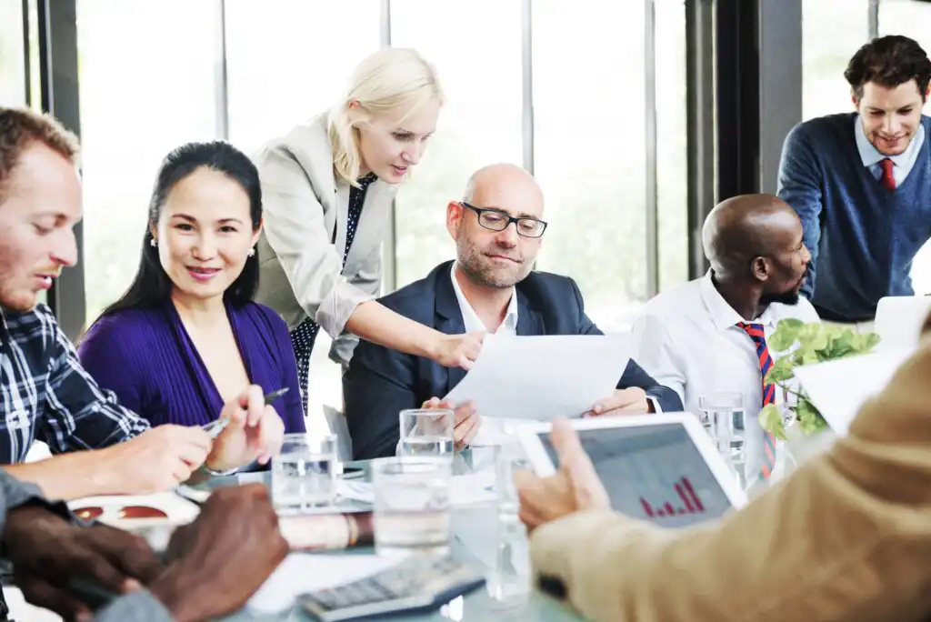 A group of six professionals sit around a conference table with documents, laptops, and water glasses, collaborating and discussing work while one woman points to a paper held by a man.