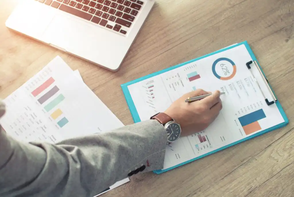 Person in a gray suit reviews financial charts and graphs on a clipboard at a wooden desk, with papers and an open laptop nearby. The graphs display data and a circular chart showing 62%.
