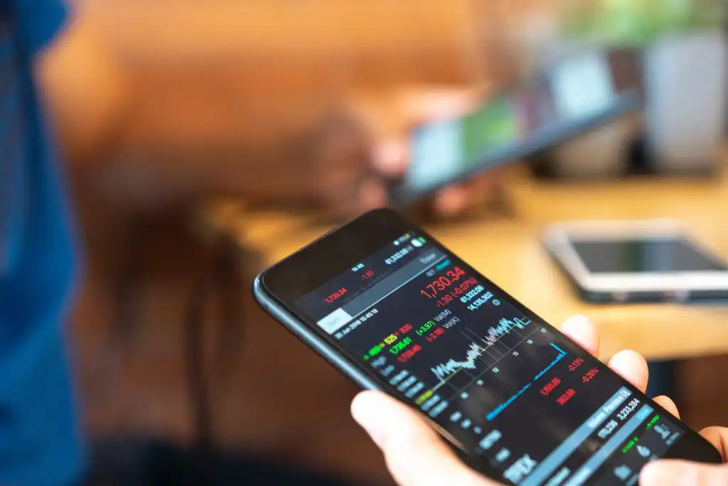 A person holds a smartphone displaying a stock market app with charts and prices, while another blurred figure uses a mobile device in the background. A table and another phone are also visible.