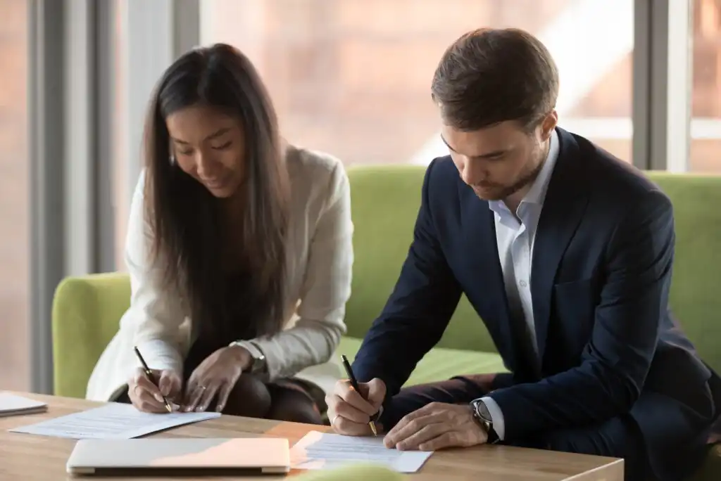 Two people sitting on a green couch at a table, both wearing business attire and signing documents with pens. Sunlight comes through the windows behind them.