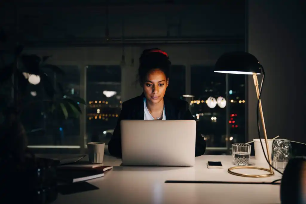 A woman sits at a desk working on a laptop at night, illuminated by a desk lamp. The room is dimly lit, and city lights can be seen through the window behind her. A notebook, phone, cup, and glass are on the desk.