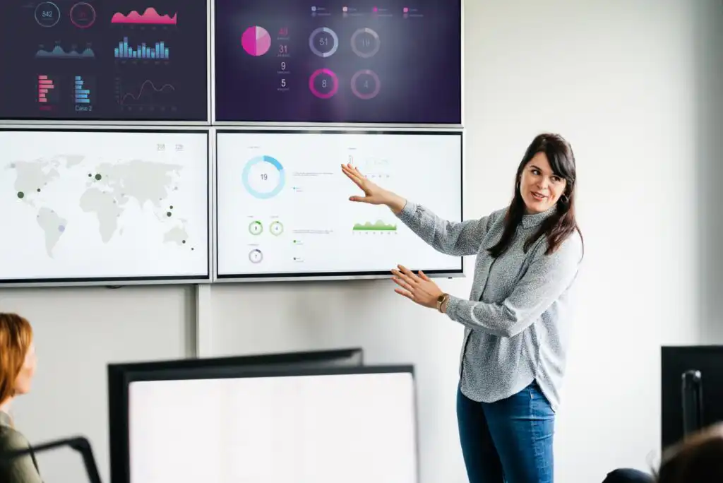 A woman stands and gestures at data visualizations on large screens, presenting information to colleagues in a modern office setting. Graphs, charts, and a world map are displayed on the screens.