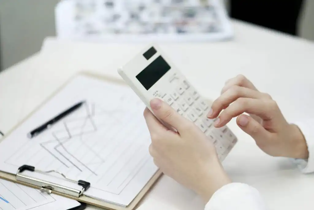 A person uses a white calculator while reviewing a clipboard with charts and graphs, and a pen rests nearby on a white desk.