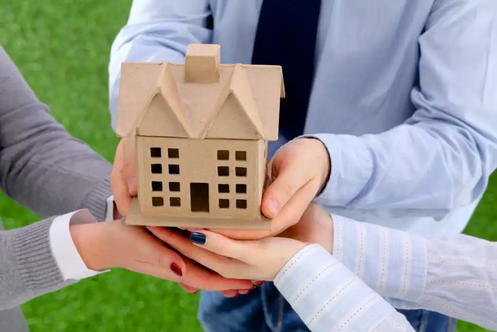 Four people hold a small cardboard model of a house together, symbolizing teamwork or shared ownership, against a background of green grass.
