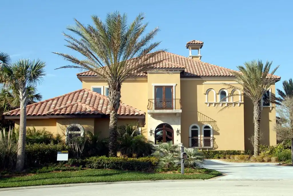 A two-story Mediterranean-style house with beige stucco walls, red-tiled roof, arched windows, and balconies, surrounded by palm trees and lush landscaping under a clear blue sky.