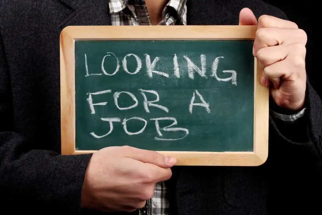 A person in a dark jacket holds a small chalkboard with the words LOOKING FOR A JOB written in white chalk.
