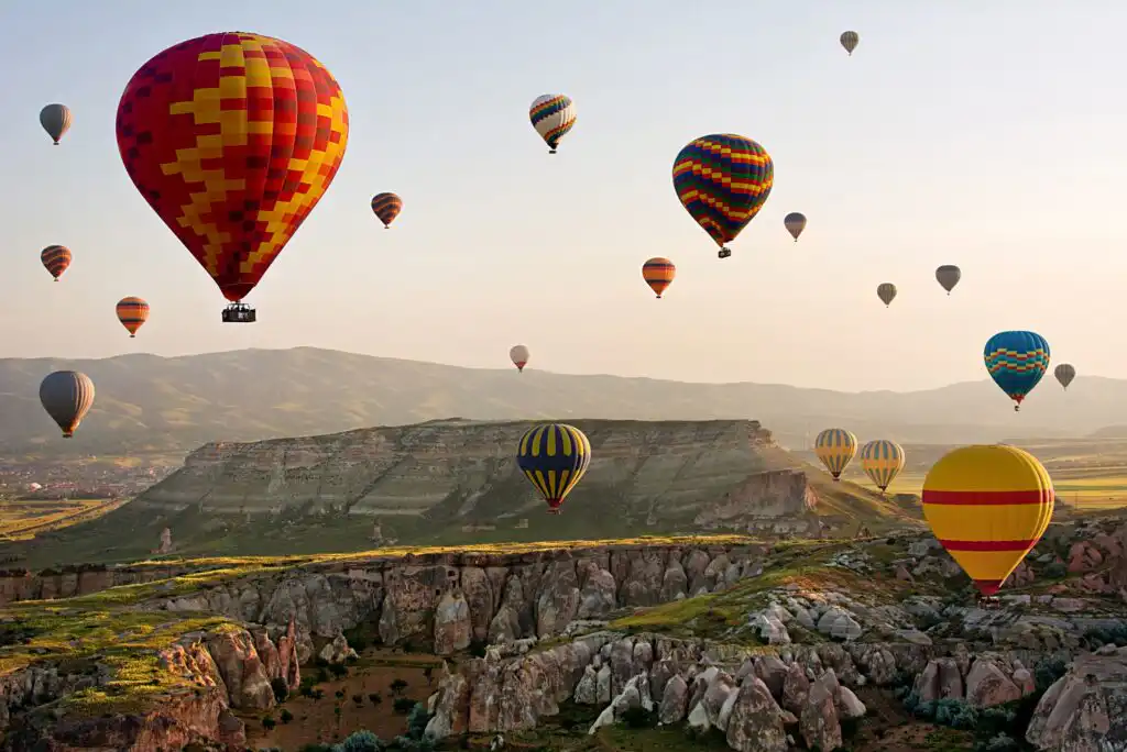 Colorful hot air balloons float above a rocky landscape with cliffs and valleys at sunrise, with distant mountains under a clear sky.