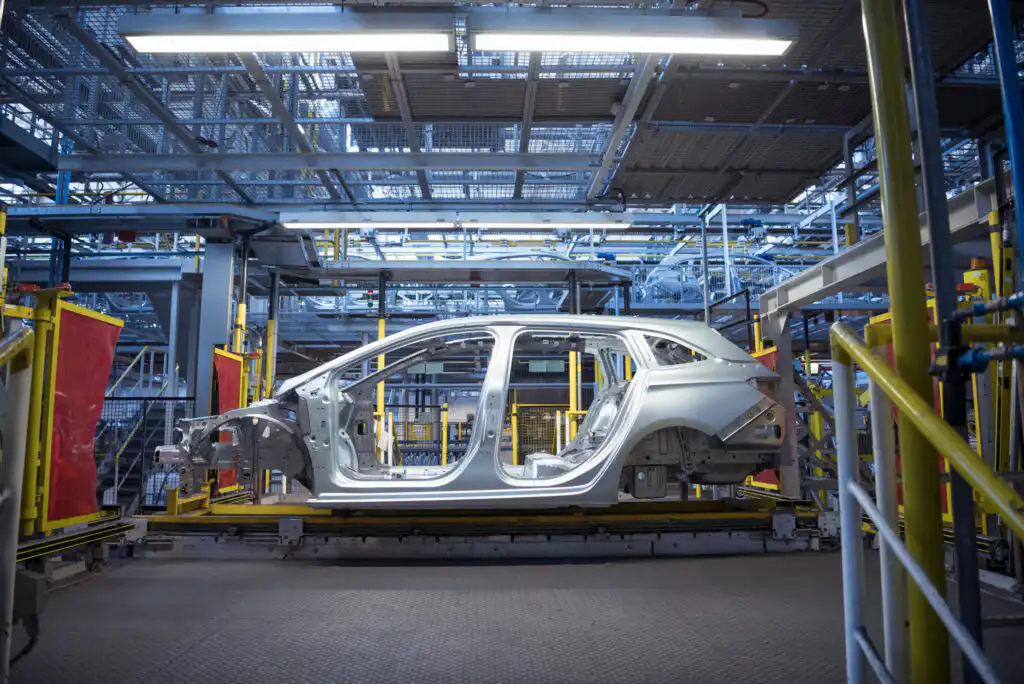 A silver car frame sits on an assembly line inside a modern, brightly lit automotive factory, surrounded by metal structures and machinery.