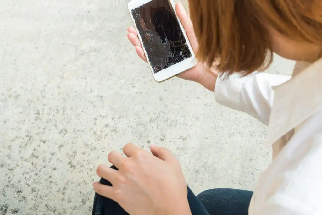 A person with light brown hair, wearing a white shirt, looks down at a smartphone with a cracked screen against a light-colored, textured surface.