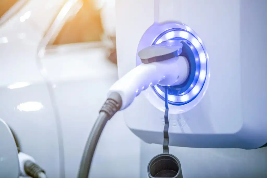 A close-up of an electric vehicle charging plug connected to a white charging station with blue LED lights, with a blurred white car in the background.