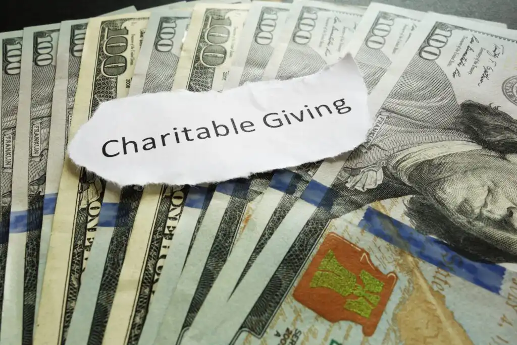 A torn piece of paper with the words Charitable Giving lies on top of a spread of U.S. hundred dollar bills.