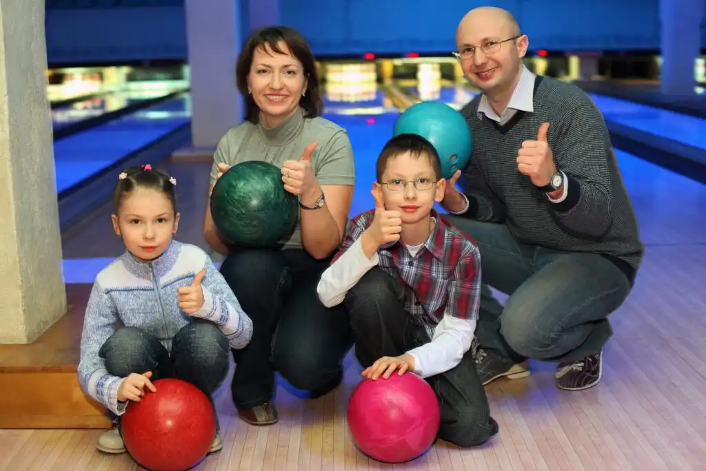 A family of four at a bowling alley smiles and gives thumbs up. The parents stand behind their son and daughter, each holding a bowling ball. The bowling lanes and pins are visible in the background.