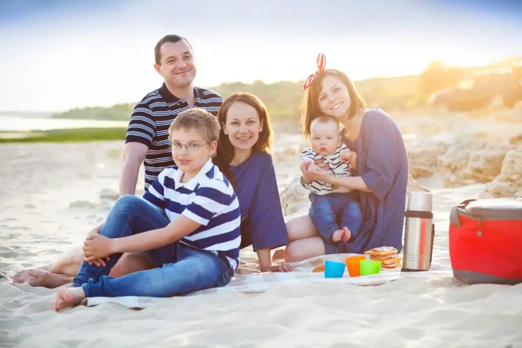 A family of five, including two adults, two children, and a baby, sits on a blanket at a sandy beach with picnic items, smiling and enjoying a sunny day.
