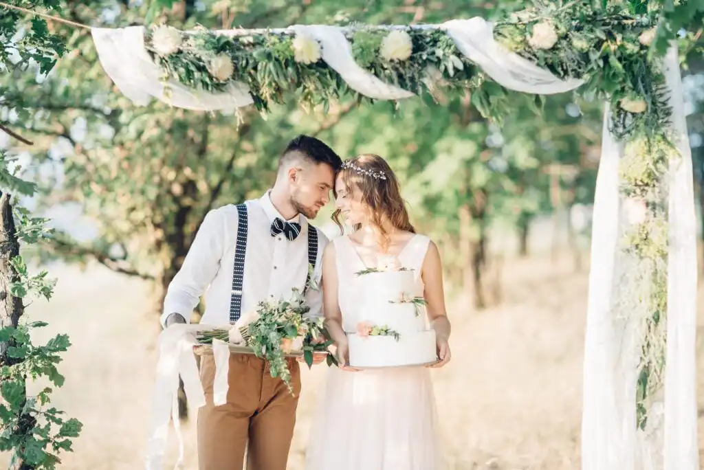 A bride and groom stand under a floral arch outdoors, smiling and touching foreheads. The groom holds a bouquet, and the bride holds a white cake decorated with greenery. Sunlight filters through the trees.