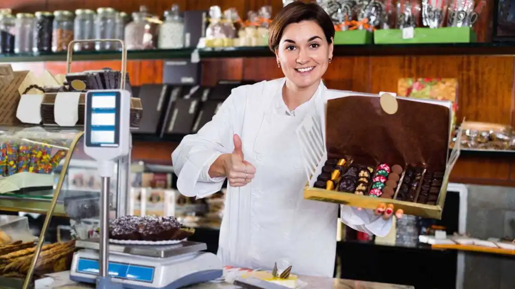 A smiling woman in a white chef coat stands behind a counter in a candy shop, holding an open box of assorted chocolates and giving a thumbs up. Shelves with jars and sweets are visible in the background.