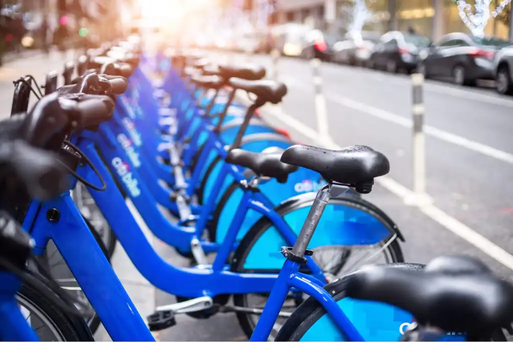 A row of blue rental bicycles is lined up at a docking station along a city street, with cars and buildings visible in the background. The photo is taken at an angle with a bright light in the distance.