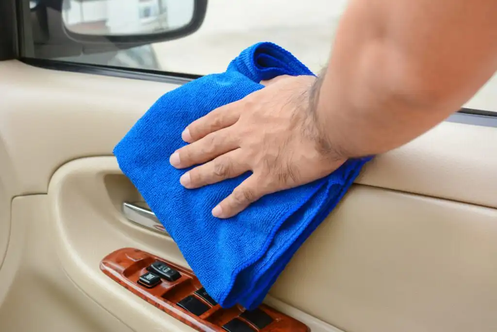 A person’s hand using a blue microfiber cloth to clean the interior door panel and controls of a beige car.