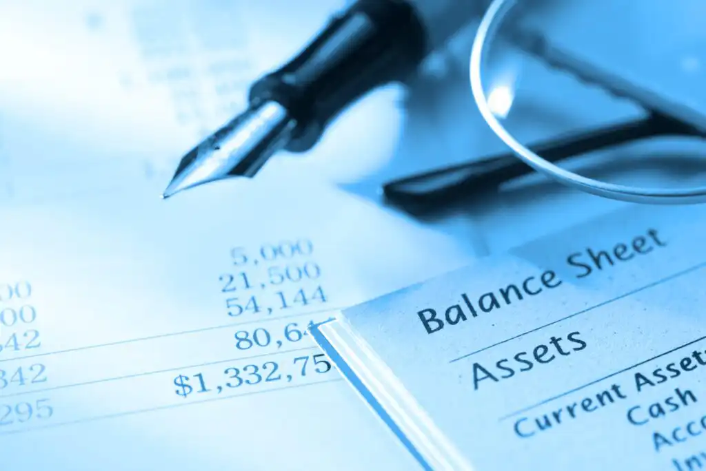 Close-up of financial documents showing numbers, a balance sheet labeled Assets, a fountain pen, and part of a pair of glasses, all tinted blue to create a professional business atmosphere.