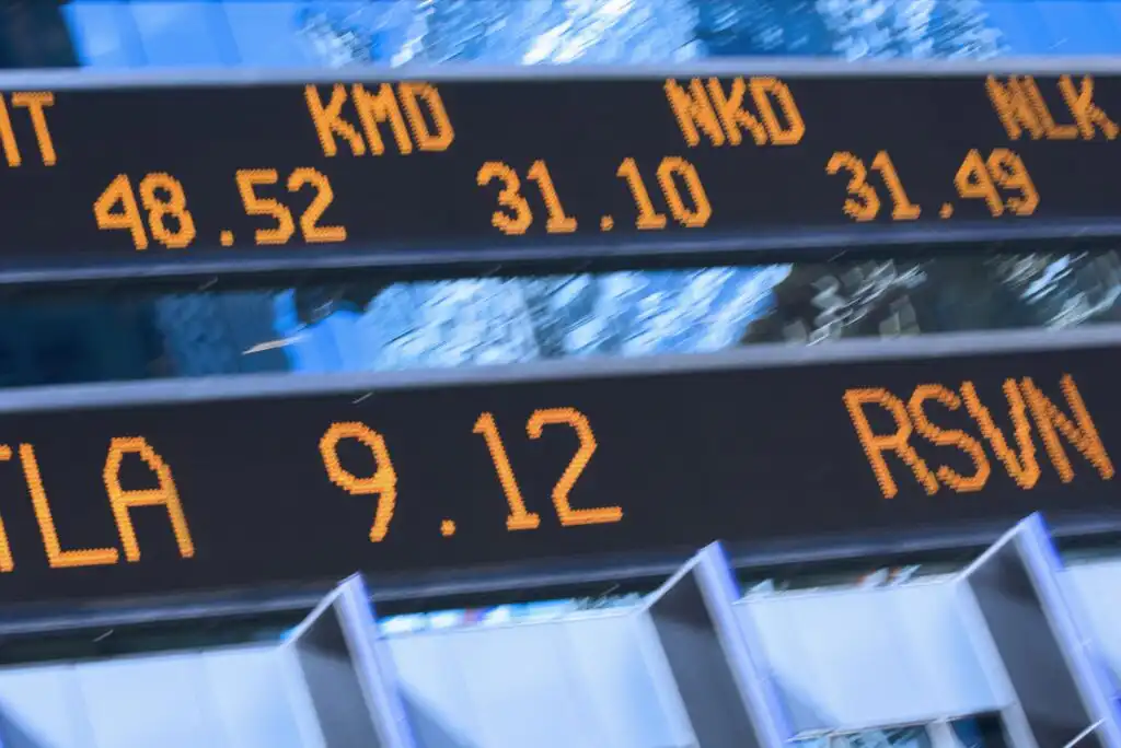 Digital stock ticker display showing stock symbols and prices in bright orange numbers against a blurred background of a glass building. Symbols include KMD, NKD, GLA, and RSVN.
