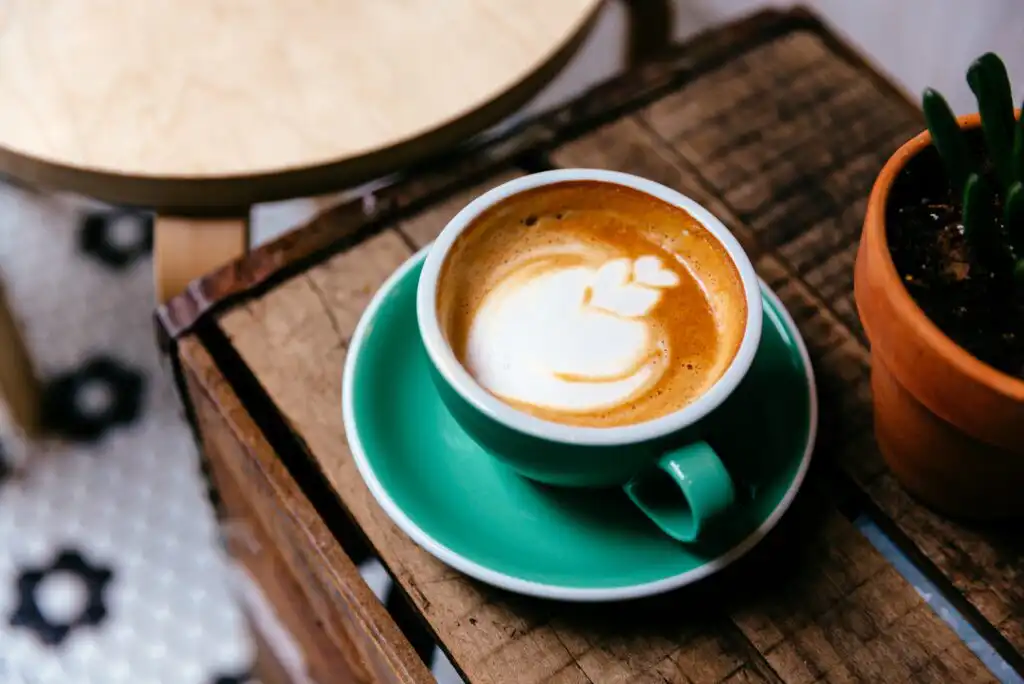 A cup of cappuccino with latte art in a green cup and saucer sits on a rustic wooden table next to a small potted cactus, with a round table visible in the background.