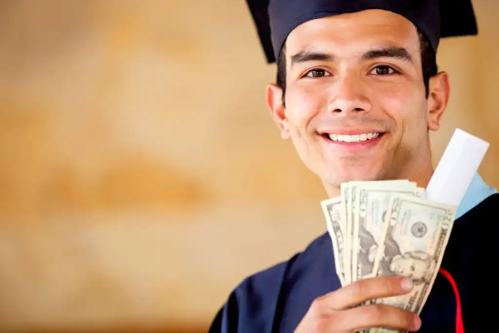 A smiling graduate in a cap and gown holds a diploma and several U.S. dollar bills in one hand, standing against a blurred, warm-toned background.