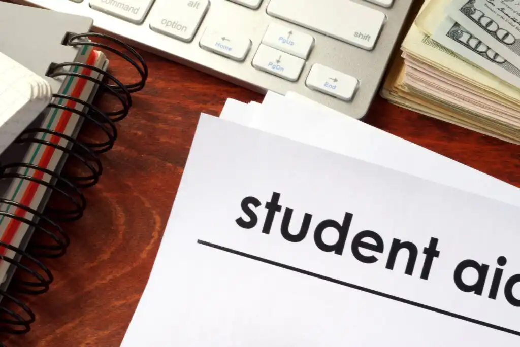 A document labeled student aid sits on a wooden desk next to a spiral notebook, a computer keyboard, and a stack of cash.