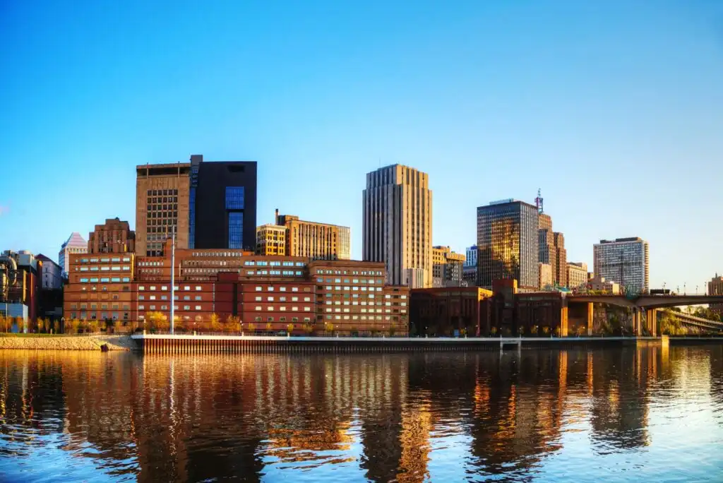 City skyline with modern and historic buildings reflecting on a calm river under a clear blue sky at sunset.