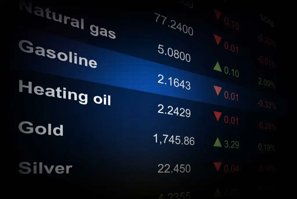 A digital screen displays commodity prices for natural gas, gasoline, heating oil, gold, and silver, with values and red or green arrows indicating price changes. Gasoline is highlighted with a blue background.