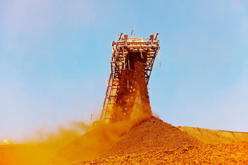 A large industrial conveyor releases reddish-brown ore onto a growing pile, with dust rising into the air against a clear blue sky at a mining site.