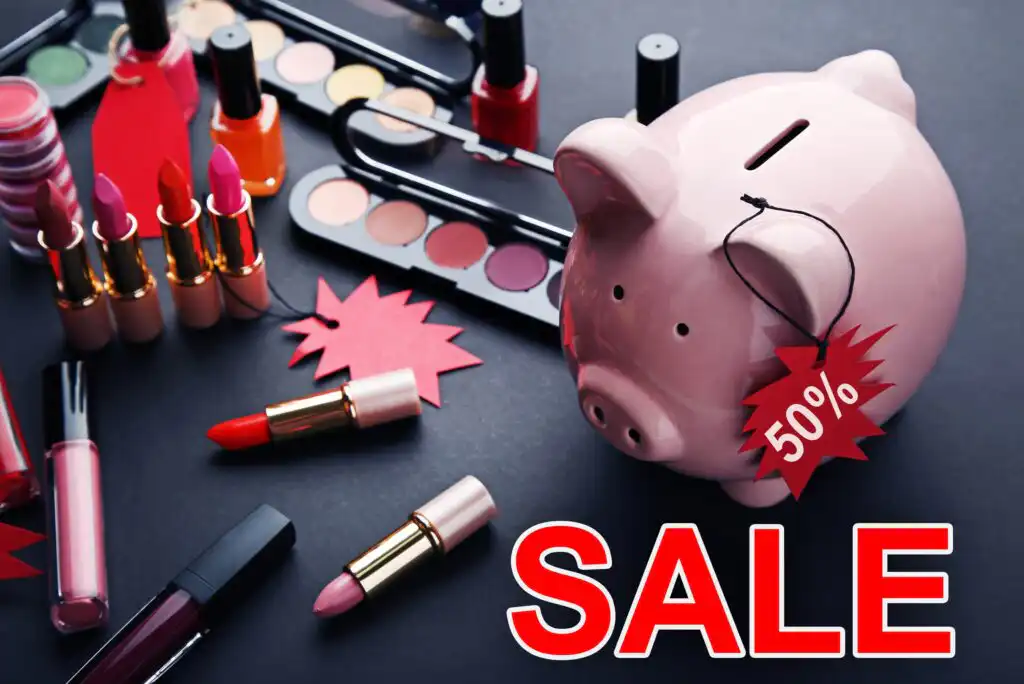 A pink piggy bank with a red 50% sale tag surrounded by various makeup products, such as lipsticks and eyeshadow palettes, on a black surface. The word SALE is prominently displayed in bold red letters.