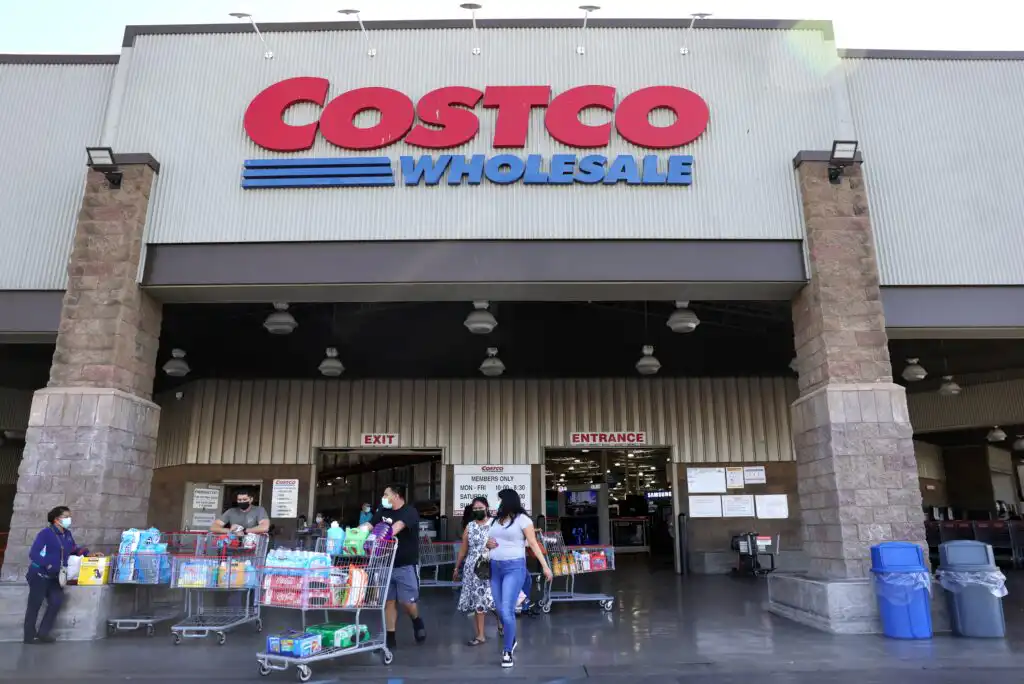 People wearing masks push carts filled with groceries outside a Costco Wholesale store. The large Costco sign is visible above the entrance, and several shoppers are entering and exiting the warehouse.