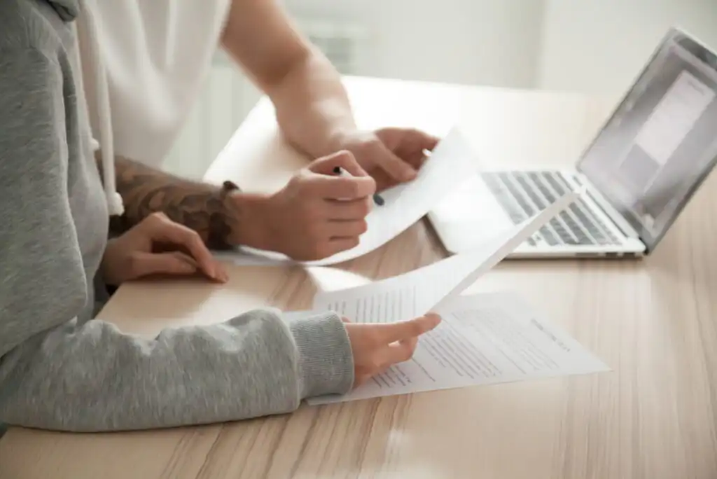 Two people sit at a table reviewing documents together, one holding papers and the other writing, with an open laptop nearby.