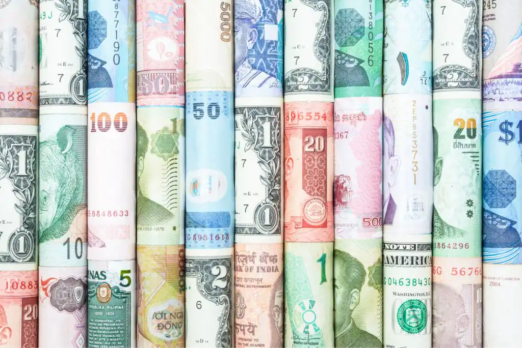 Rolled-up banknotes from various countries and currencies are arranged vertically side by side, displaying a colorful mix of numbers, symbols, and faces.