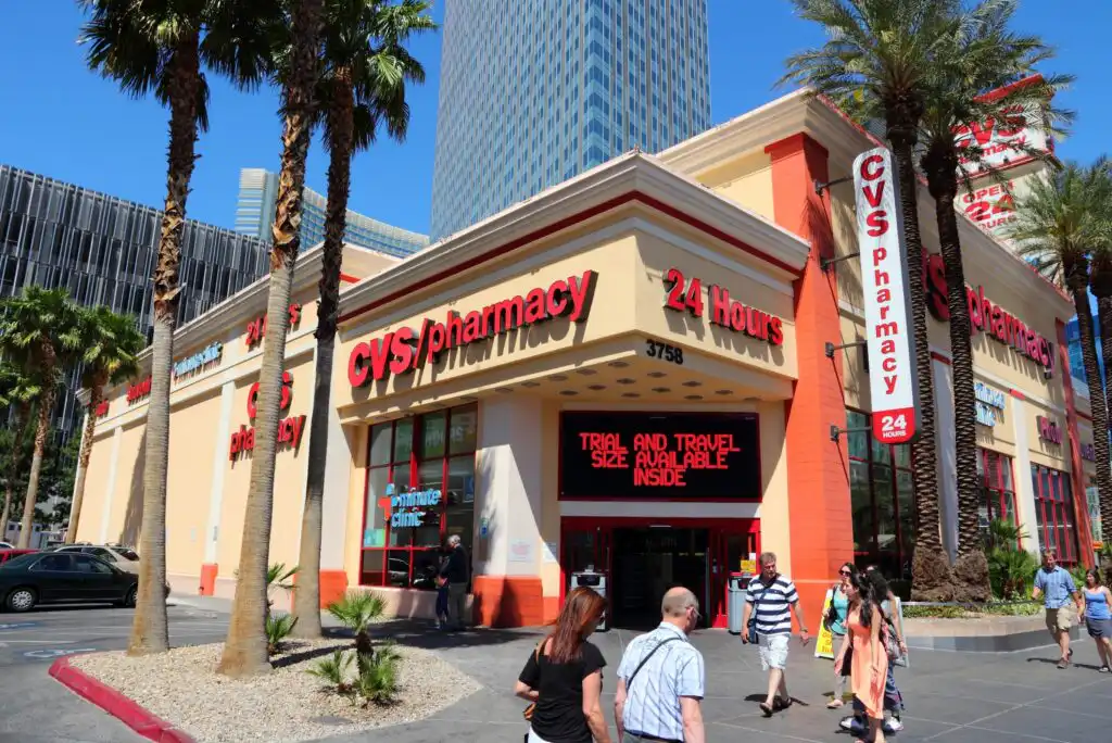 A CVS Pharmacy store with large red signage, open 24 hours, located on a city street. Several people are walking near the entrance, and tall palm trees line the sidewalk.