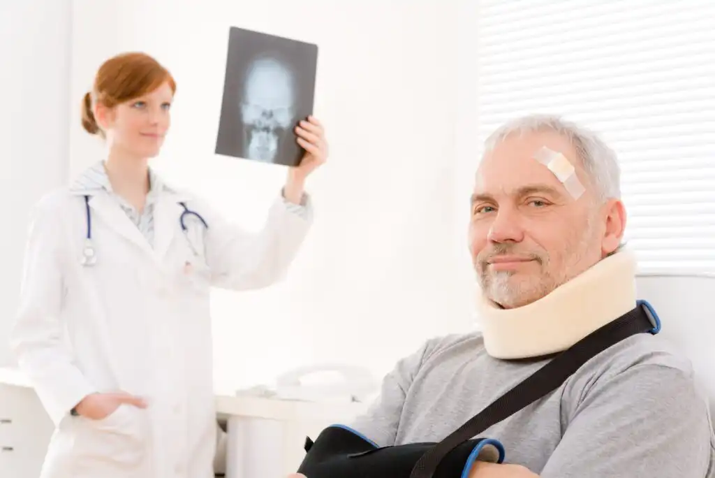 A man with a bandage on his forehead, wearing a neck brace and arm sling, sits and smiles. A doctor in a white coat holds up an X-ray image of a skull in the background.