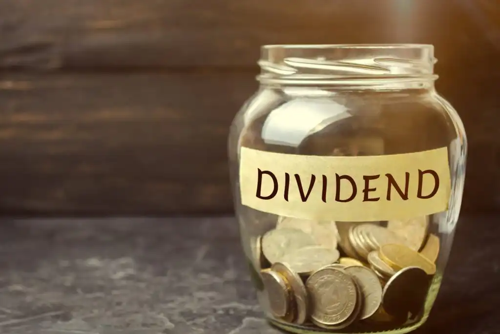 A glass jar labeled DIVIDEND is filled with assorted coins and placed on a dark surface, with a blurred dark background and light shining from the top right corner.