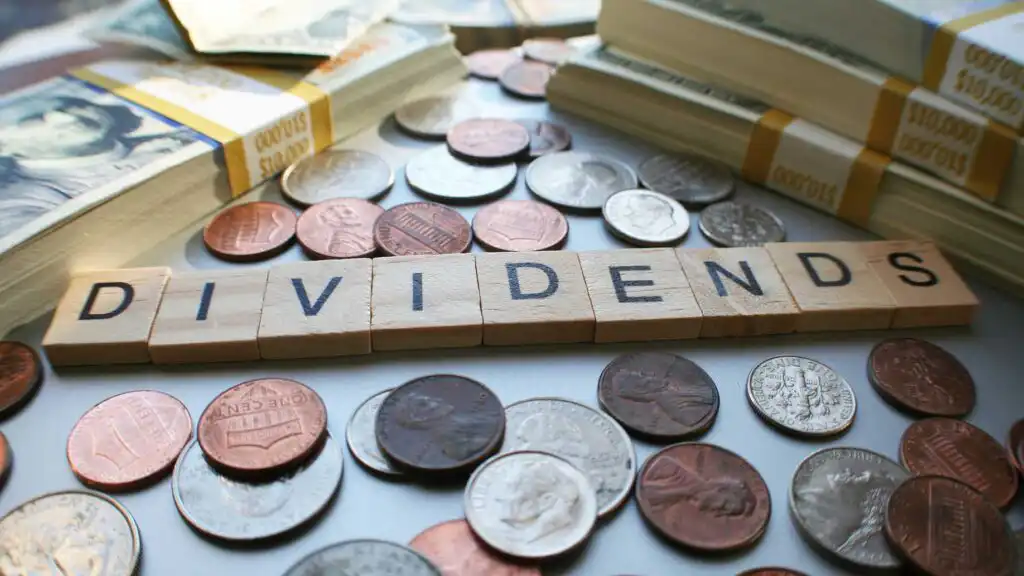 Scrabble tiles spell DIVIDENDS surrounded by scattered coins and stacks of U.S. dollar bills.