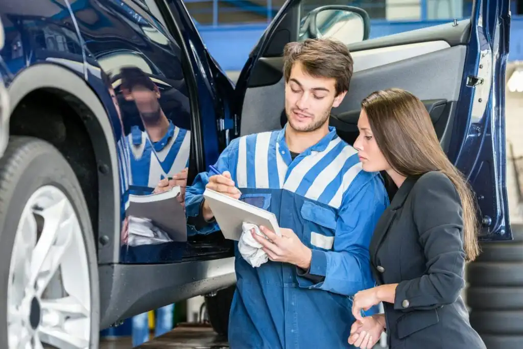 A mechanic in blue overalls explains repair details on a clipboard to a woman in business attire beside a raised car in an auto repair shop.