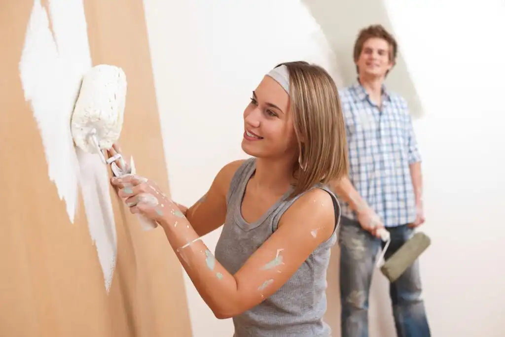 A woman smiles while painting a wall white with a roller, paint on her arms and clothes. A man in the background, also holding a roller, watches her and smiles. Both are dressed casually and appear to be renovating.