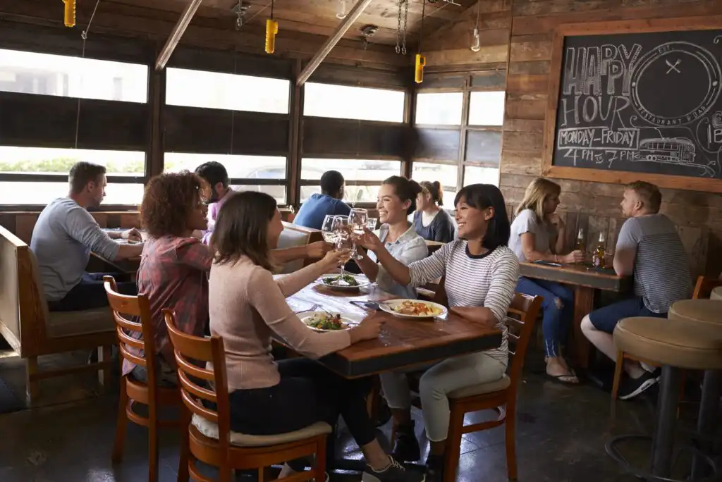 A group of friends sit at a wooden table in a rustic restaurant, raising their glasses in a toast. Other diners are seated at nearby tables. The atmosphere is lively, with a chalkboard advertising happy hour on the wall.