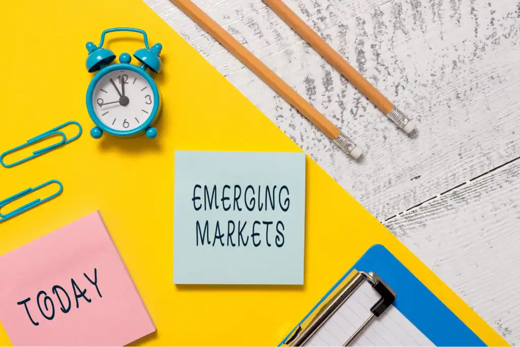 A blue alarm clock, two pencils, paper clips, and sticky notes reading TODAY and EMERGING MARKETS are arranged on a bright yellow and white desk next to a clipboard.