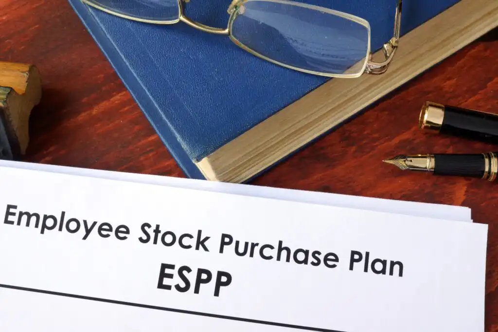A document titled Employee Stock Purchase Plan (ESPP) on a wooden desk, with a blue book, a pair of eyeglasses, and a fountain pen beside it.