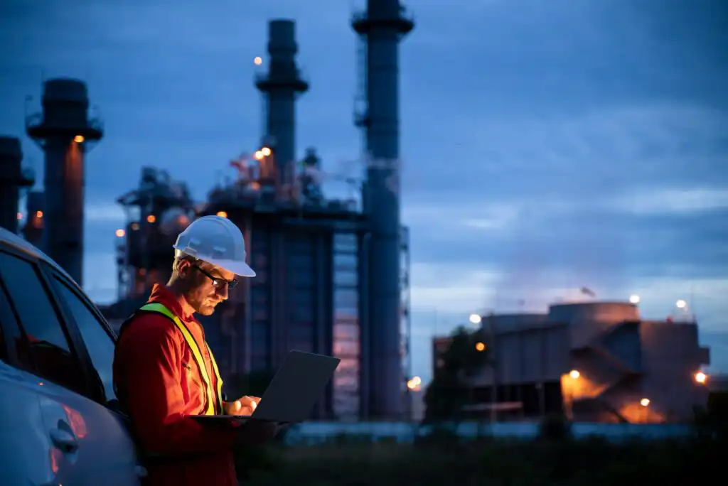 A worker in a hard hat and safety vest stands by a vehicle at dusk, using a laptop. Industrial structures and bright lights are visible in the background.