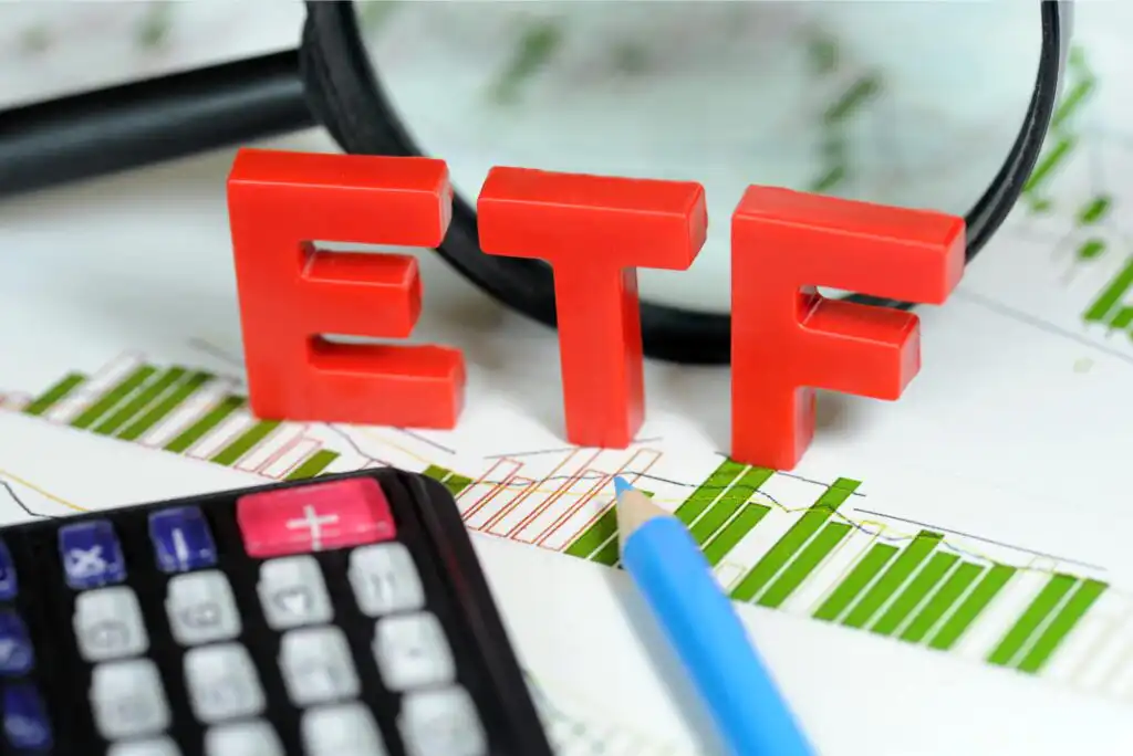 Red letters spelling ETF stand on financial charts with green bars. A calculator, blue pencil, and magnifying glass are also visible, symbolizing investment and stock market analysis.