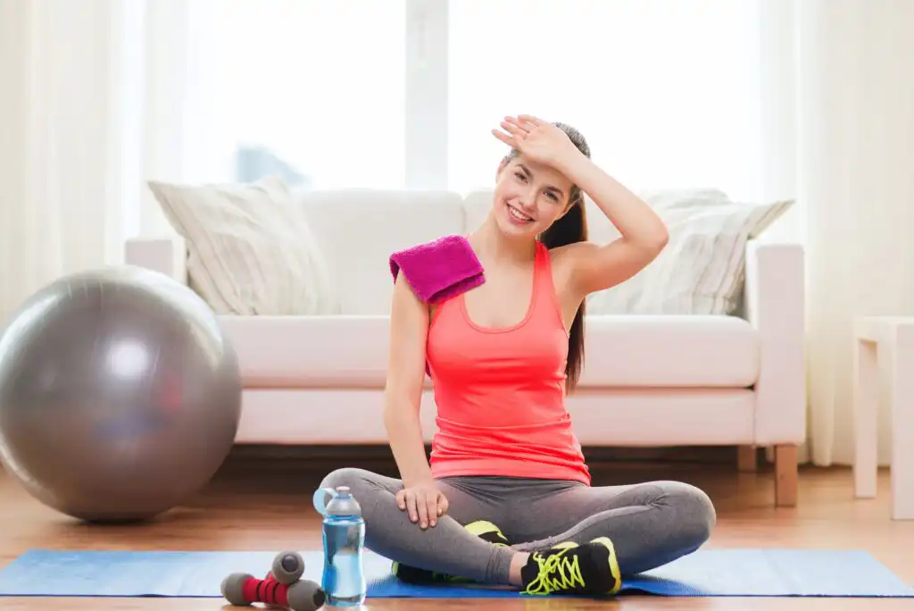 A smiling woman in workout clothes sits on a yoga mat indoors, wiping her forehead. A pink towel is on her shoulder, with dumbbells, a water bottle, and an exercise ball nearby. A couch is in the background.