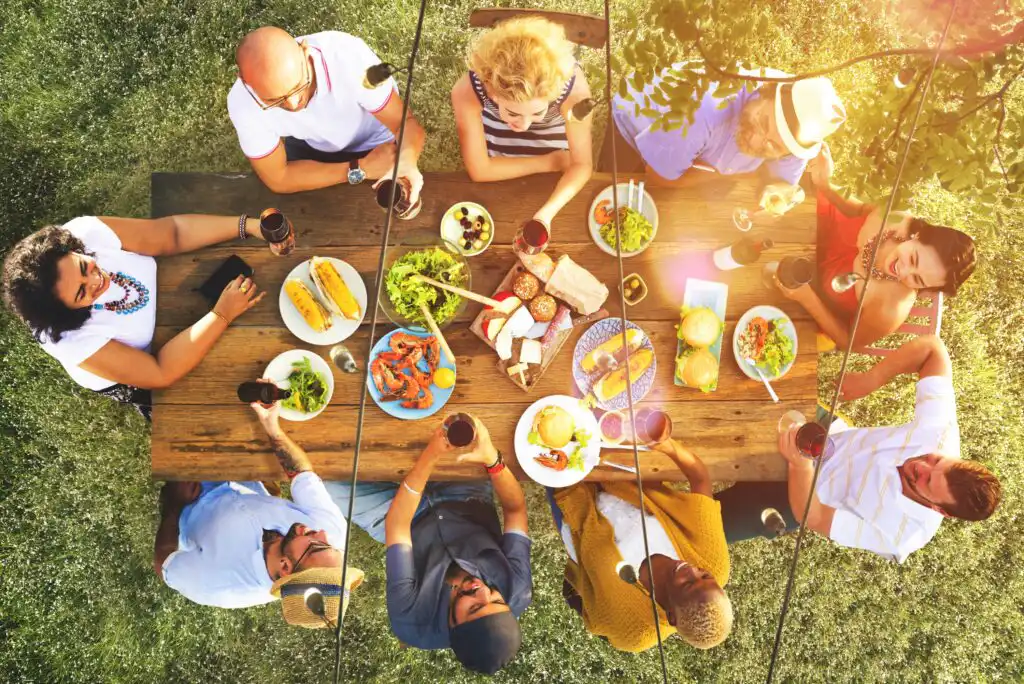 A group of people sit around a wooden table outdoors, sharing a meal and drinks. The table is filled with various foods like salads, bread, and grilled items. The scene is bright and lively, set on a grassy lawn.