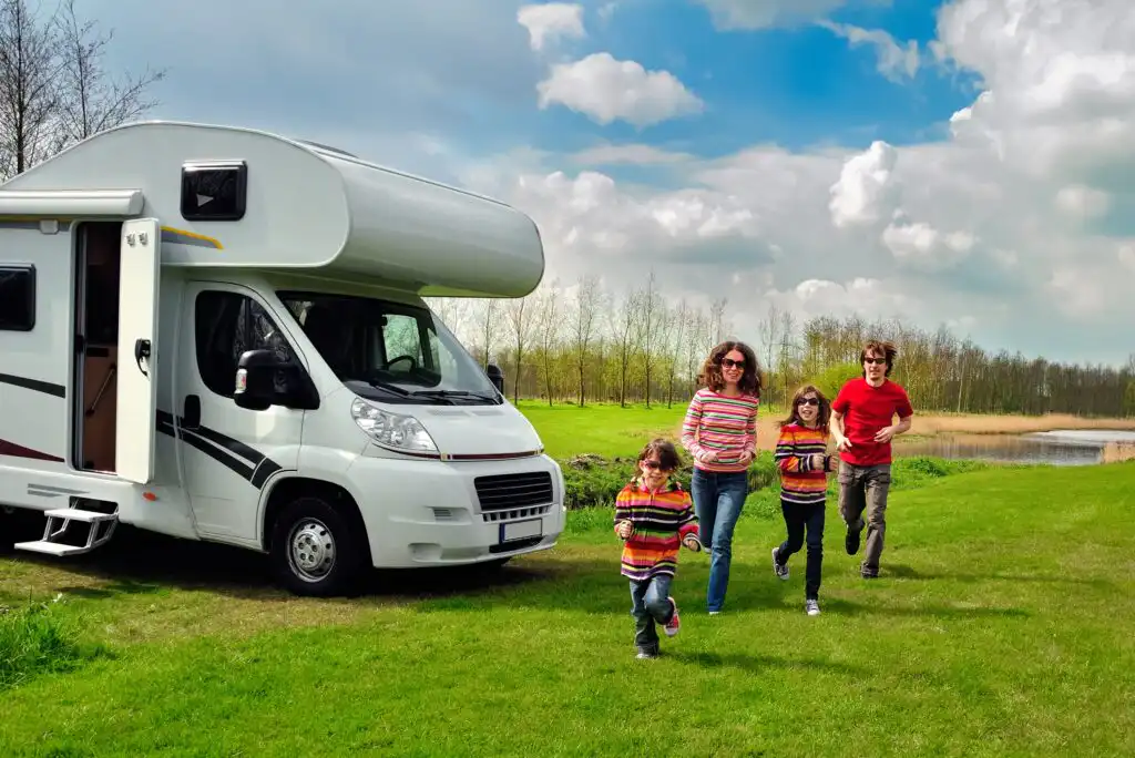 A family of four, wearing colorful sweaters, runs joyfully on green grass near a white camper van parked beside a pond, with blue sky and clouds overhead.