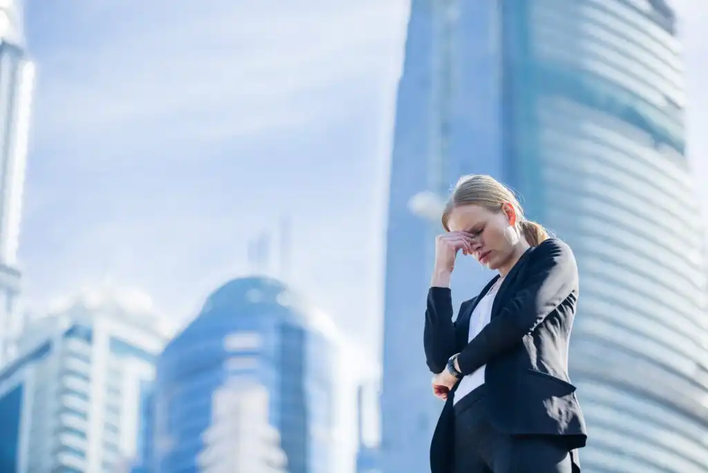 A woman in business attire stands outside in a city, appearing stressed or tired, with her hand on her forehead and eyes closed. Skyscrapers with reflective glass are visible in the background.