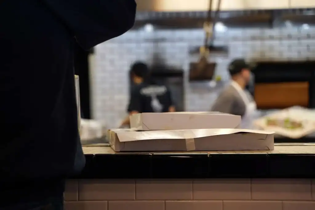 Two closed pizza boxes sit on a tiled counter in a restaurant kitchen, with two blurred people working in the background near a brick oven.